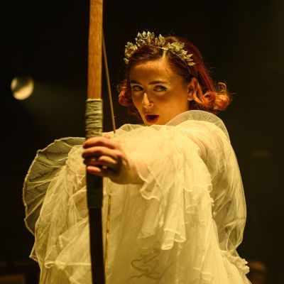 Opera student wearing a long tulle dress holding a bow and arrow aiming for the centre of the screen
